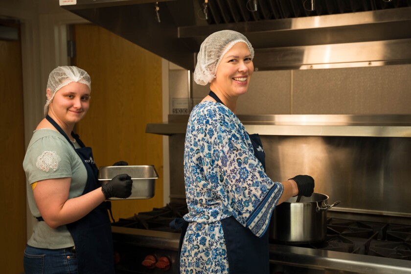 Rees, wearing a blue floral shirt and hairnet, stirs a pot while a student in a green shirt and hairband holds a tray. Both stand in front of an industrial stove. 