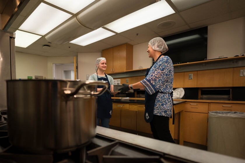 Rees, wearing a blue floral shirt and hairband, carries a tray as she talks to a student in a green shirt and hairnet behind a pot on an industrial stove. 