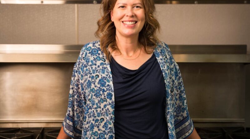 A woman in a black shirt and floral jacket stands proudly in front of an industrial stove.