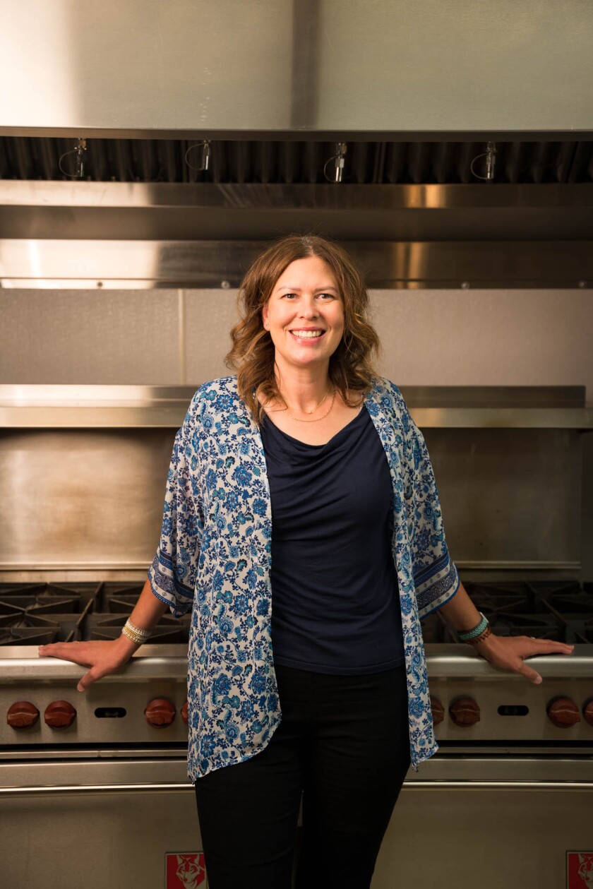 A woman in a black shirt and floral jacket stands proudly in front of an industrial stove. 